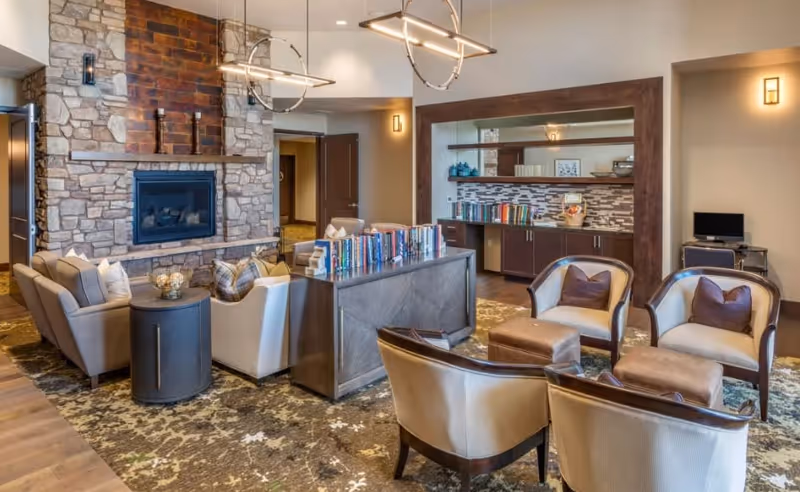 A cozy living room area in Ansel Park Independent Living featuring a stone fireplace with a wooden mantel, surrounded by beige armchairs and small ottomans. A wooden console table with books is placed in the center, and a built-in cabinet with a decorative backsplash and mirror is visible in the background. Modern pendant lights hang from the ceiling, and a patterned rug covers the floor.