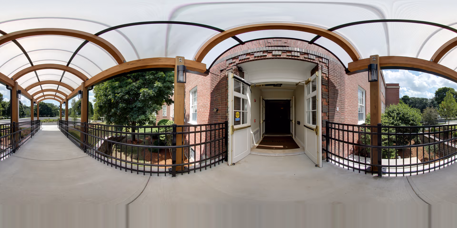 Covered walkway with wooden arches and a translucent roof leading to an open double door entrance of a brick building, surrounded by greenery and trees on a sunny day.