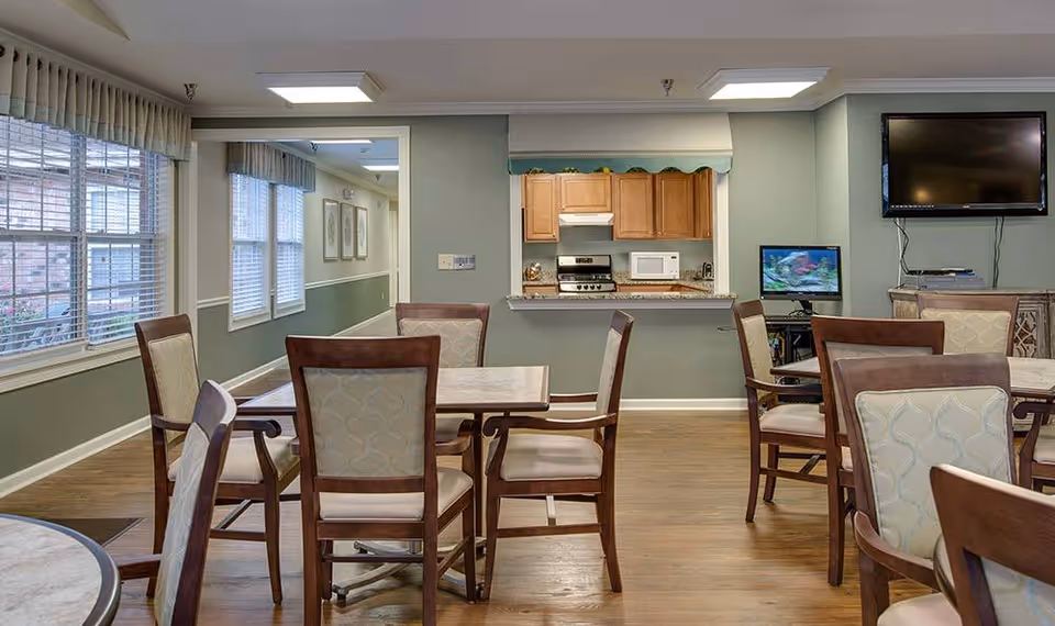 Communal dining area with wooden tables and upholstered chairs, a serving kitchen pass-through, windows on the left, and a wall-mounted TV.