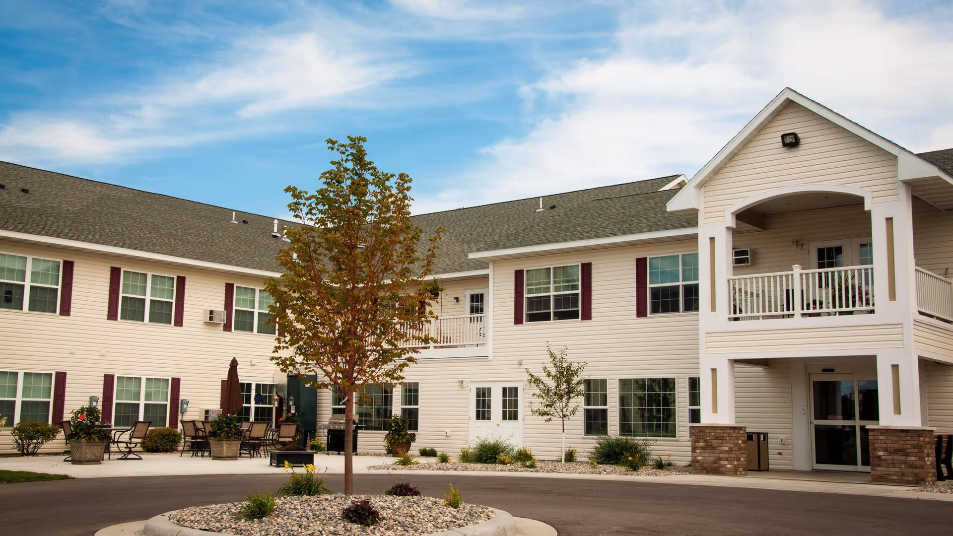 Exterior view of a two-story assisted living community building with white siding and maroon shutters. There is a small tree planted in a circular landscaped area in front of the building, outdoor seating with tables and chairs, and a covered balcony above the main entrance. The sky is partly cloudy.
