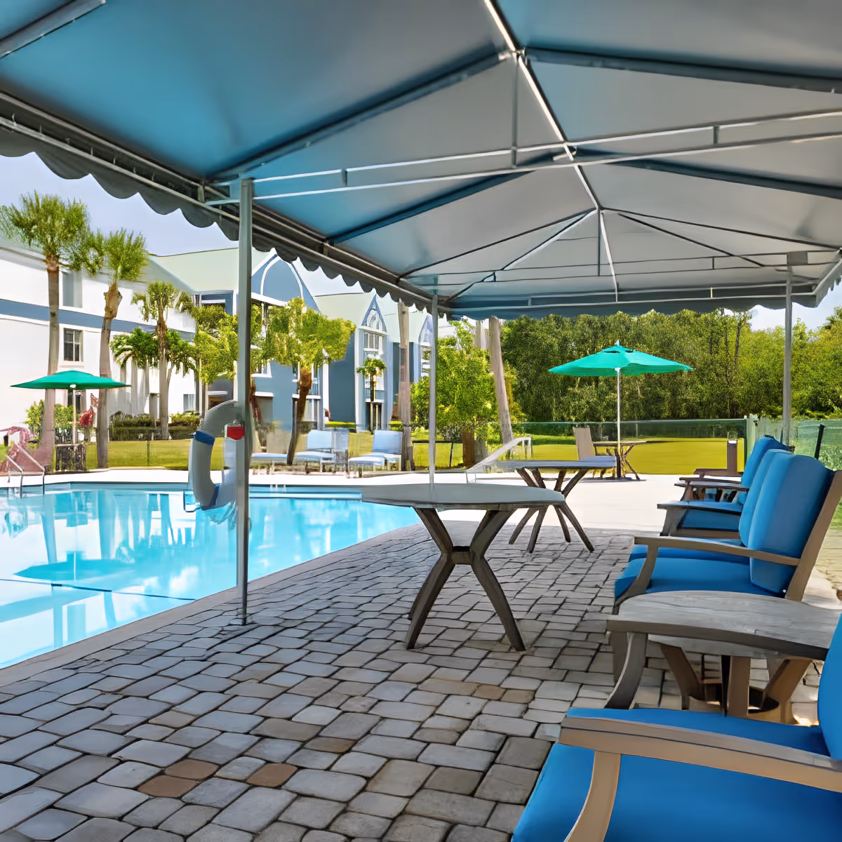 Covered poolside seating area with blue cushioned chairs, tables, umbrellas, and a swimming pool by a residential building.
