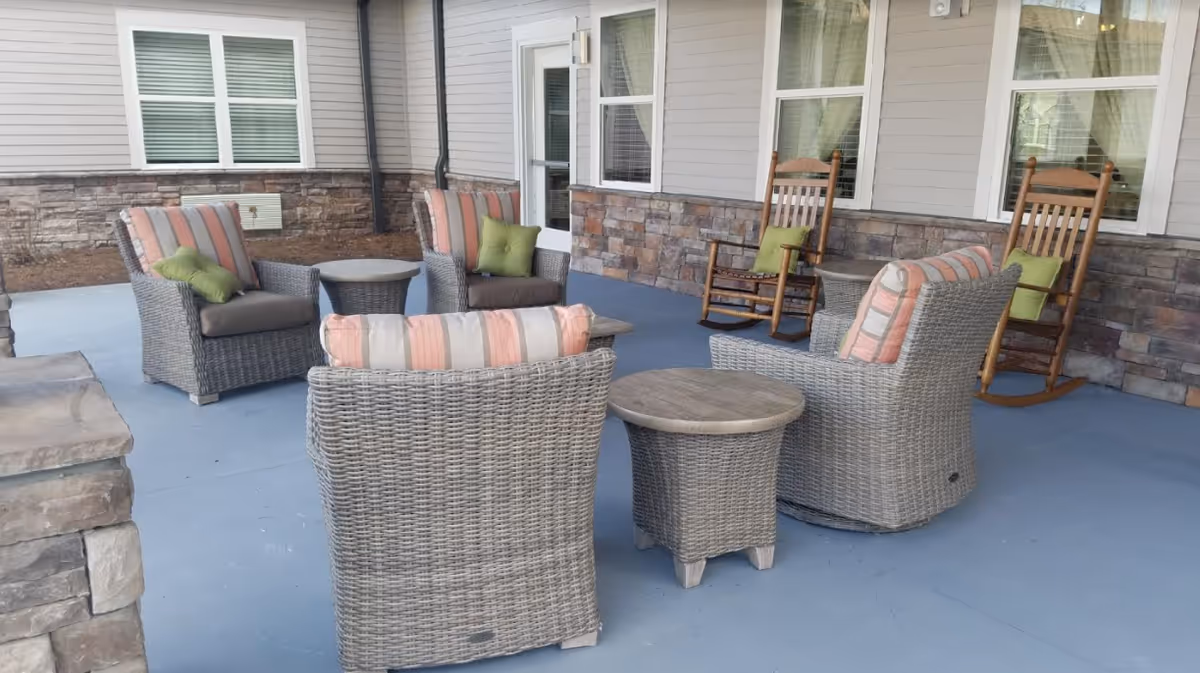 Outdoor patio area with wicker chairs and small tables arranged on a blue concrete floor. The chairs have striped cushions in shades of pink, beige, and green. Two wooden rocking chairs with green cushions are positioned near the building wall with windows and a door.