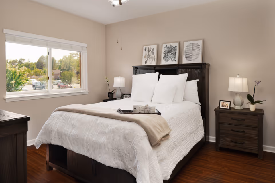A cozy bedroom with a large bed featuring white bedding and multiple pillows. The bed has a dark wooden headboard with three framed artworks above it. There are two matching dark wooden nightstands on either side of the bed, each with a lamp and decorative items including a small plant and a framed photo. A window on the left side lets in natural light, showing trees and parked cars outside. The room has beige walls and wooden flooring.