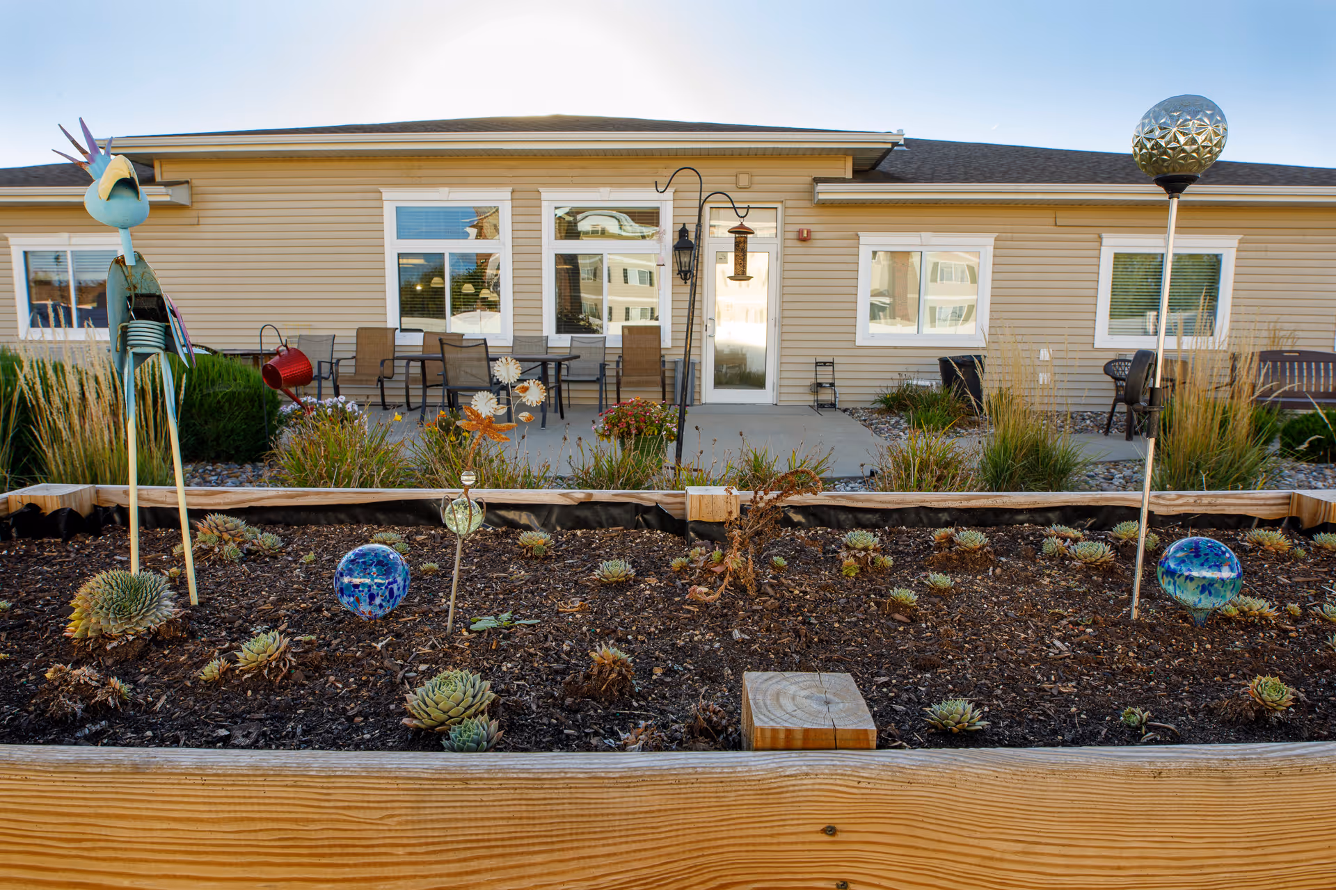 Outdoor garden area with decorative plants and ornaments in front of a beige building with windows and a door. There are patio chairs and tables on a concrete patio, along with some garden decorations including a bird sculpture and glass spheres.