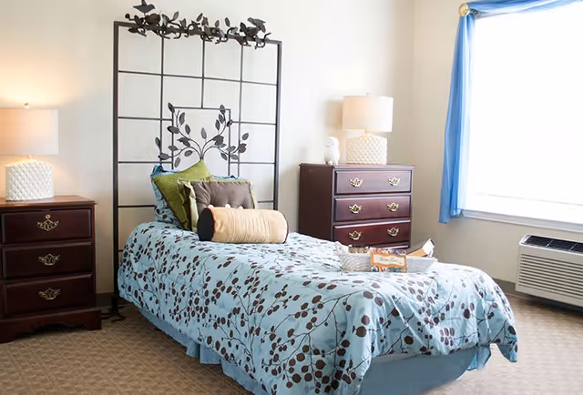 A bright bedroom with a single bed featuring a decorative metal headboard with leaf designs. The bed is covered with a light blue comforter with a brown leaf pattern and several pillows in green, brown, and beige. On either side of the bed are wooden nightstands with brass handles, each topped with a white lamp. A window with blue curtains allows natural light to fill the room, and there is an air conditioning unit beneath the window.