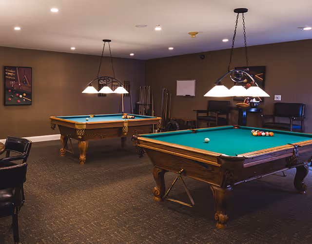 Interior room with two wooden pool tables with green felt tops, overhead hanging lights, several chairs, and pool cues against the wall. The room has brown walls and carpeted floor.