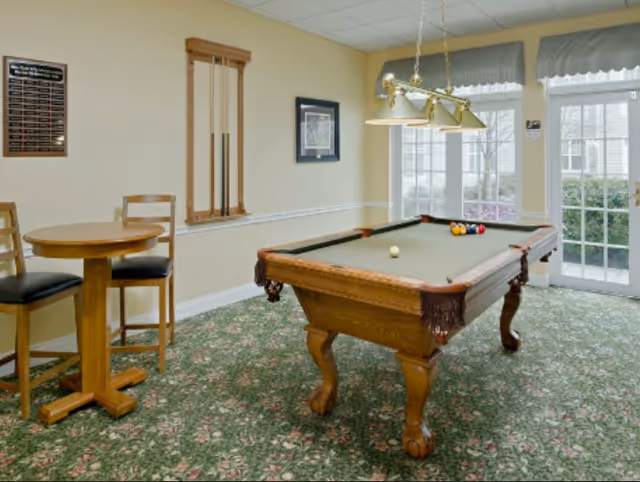 A recreational room with a wooden pool table set up for a game, a small round wooden table with two chairs, a wall-mounted pool cue rack, framed artwork on the wall, and large windows with a door leading outside. The room has floral patterned carpet and beige walls.