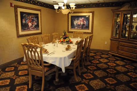 A formal dining room with a long wooden table covered with a white tablecloth, set with folded napkins, glassware, and silverware. The room has wooden chairs with decorative backs, two framed floral paintings on the walls, a chandelier overhead, and a wooden china cabinet filled with dishes. The carpet has a patterned design.