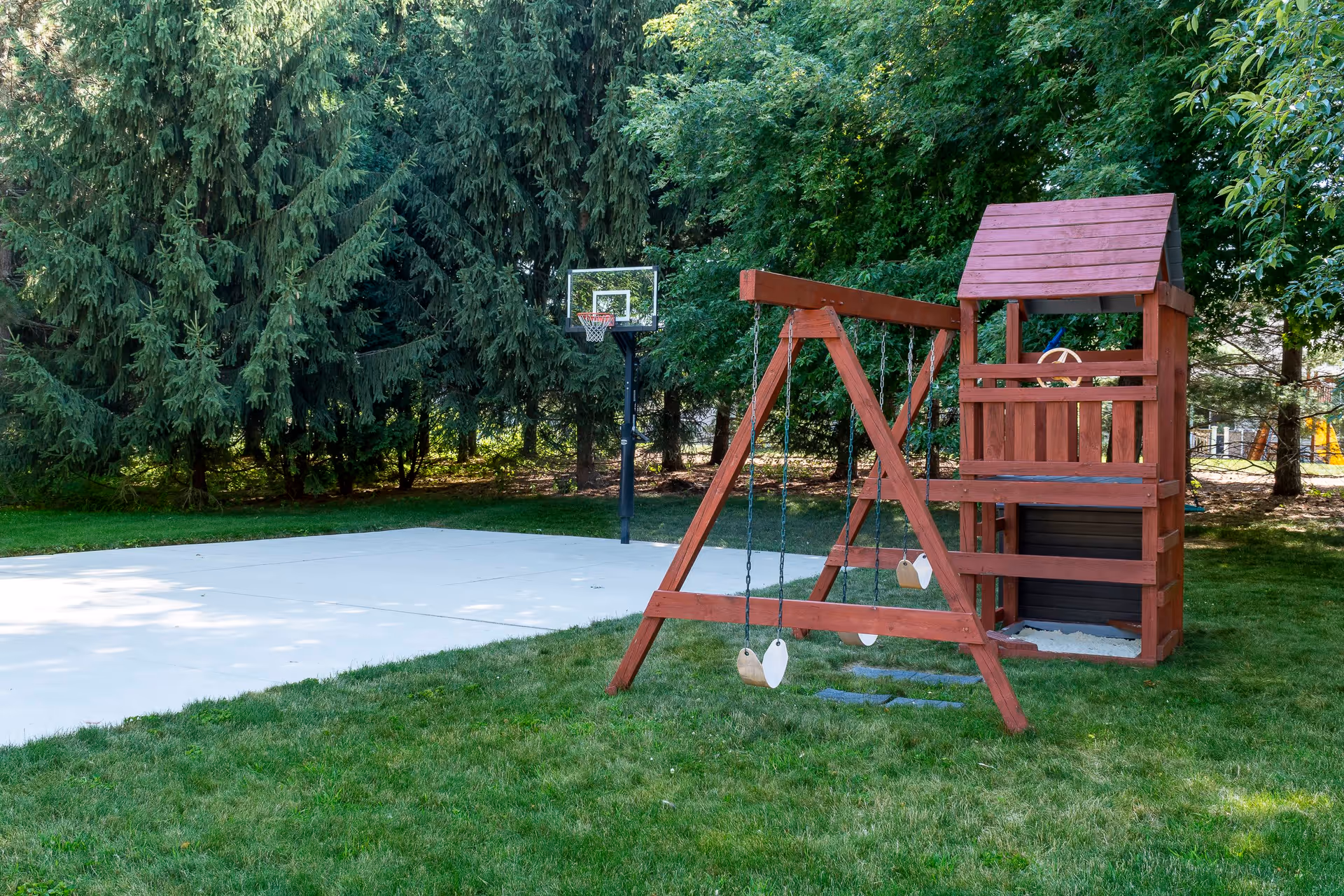 Wooden children's playset with swings beside a concrete basketball half-court in a grassy, tree-lined yard.
