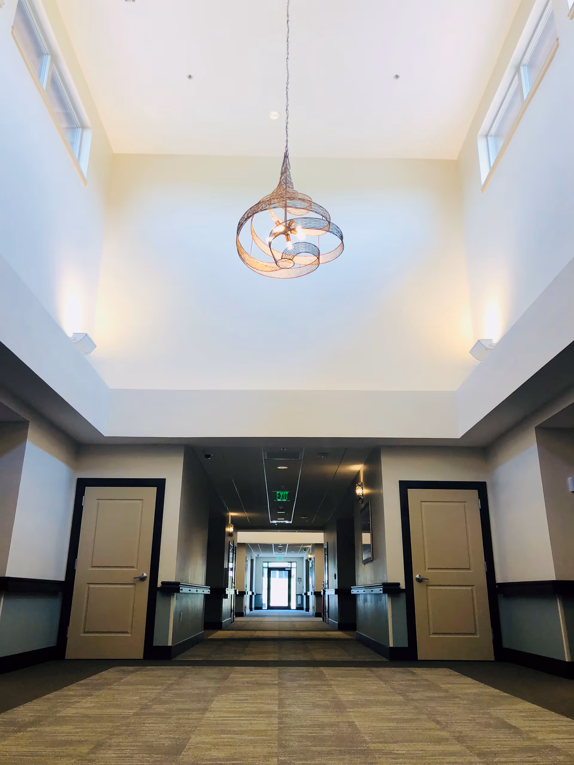 A wide interior hallway with a high ceiling and a central decorative pendant light, flanked by doors and leading to an exit.