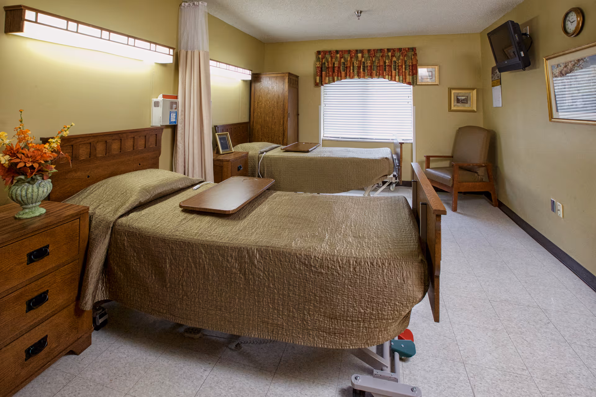 Two hospital-style beds with brown bedspreads in a senior living bedroom with wooden furniture, a chair, TV, and a window with blinds.