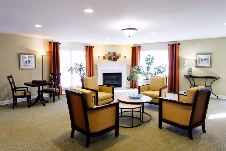 A bright and spacious living room with four beige armchairs arranged around a round coffee table. The room features a white fireplace centered on the far wall, flanked by large windows with orange curtains. There are two framed pictures on the walls, a small table with two chairs on the left, and a console table with a lamp on the right. Several green plants add a touch of nature to the space.