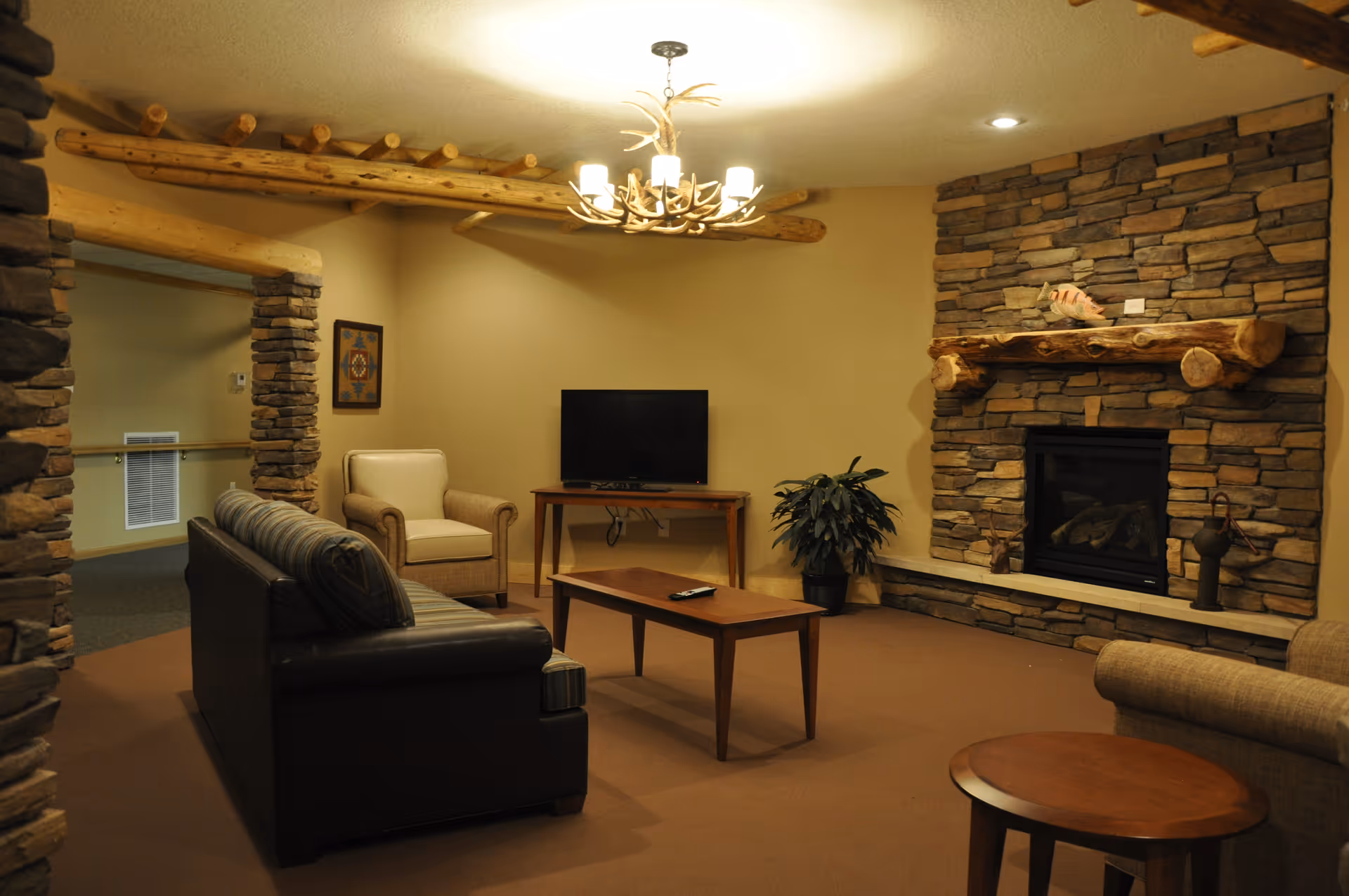 A cozy living room with a stone fireplace and wooden mantel, a flat-screen TV on a wooden table, a black leather sofa, a beige armchair, a wooden coffee table, a round wooden side table, a potted plant, and a chandelier with antler design hanging from the ceiling.