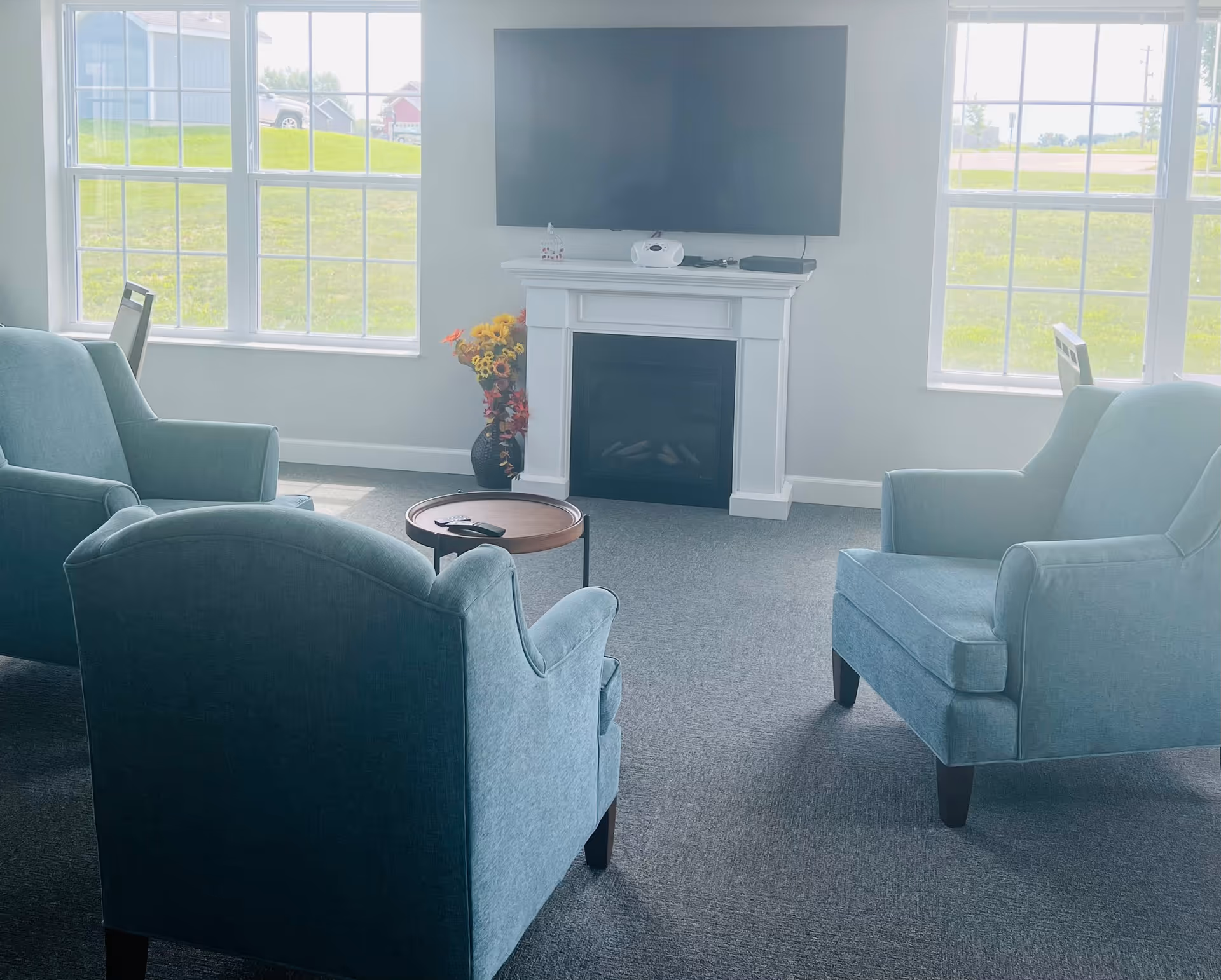 A cozy living room area with three blue upholstered armchairs arranged around a small round wooden table. A flat-screen TV is mounted above a white electric fireplace, with a vase of colorful flowers placed beside it. Large windows on either side let in natural light and show a grassy outdoor area.
