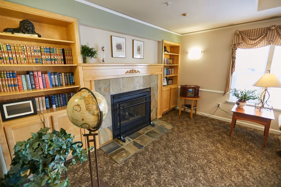 A cozy living room area featuring a fireplace with a tiled surround and wooden mantel. On either side of the fireplace are built-in wooden bookshelves filled with books and decorative items. In front of the fireplace is a globe on a stand and a potted plant. To the right, there is a small wooden table with a lamp and a potted plant next to a window with striped curtains. A small wooden side table with a stereo system is also visible.