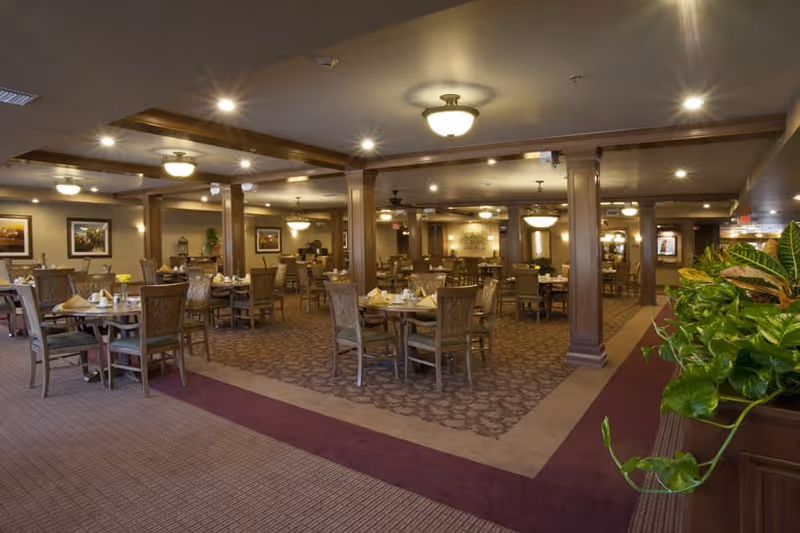 Spacious dining room with multiple round tables set with napkins and tableware, surrounded by wooden chairs. The room features carpeted floors, wooden columns, ceiling lights, and framed artwork on the walls. There are green plants on the right side near the entrance.