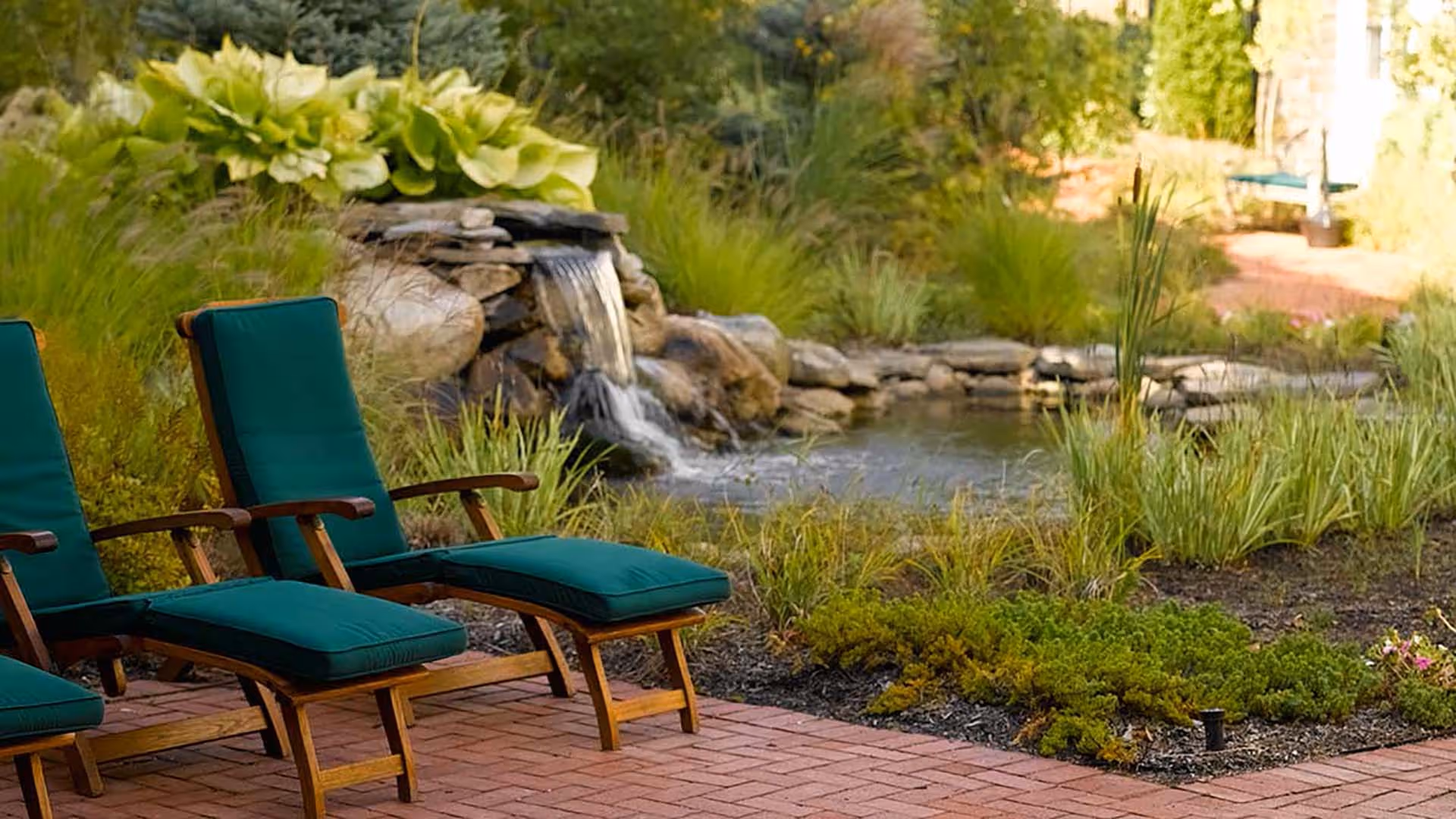 Two wooden lounge chairs with green cushions placed on a brick patio next to a small pond with a rock waterfall surrounded by lush green plants and grass.