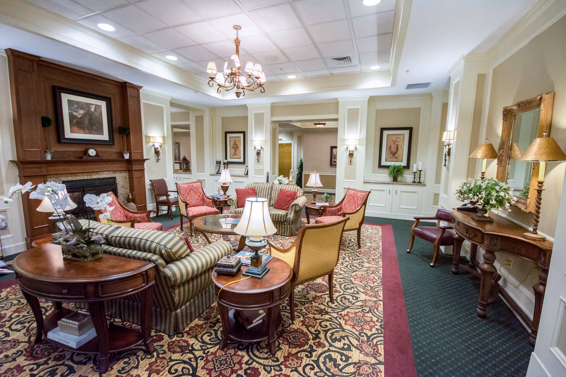 A cozy and elegant living room area with a patterned carpet, striped and red upholstered chairs, a wooden coffee table, side tables with lamps, a fireplace with a framed picture above it, wall sconces, and a chandelier hanging from the ceiling. There is also a wooden console table with a large mirror and decorative flowers.