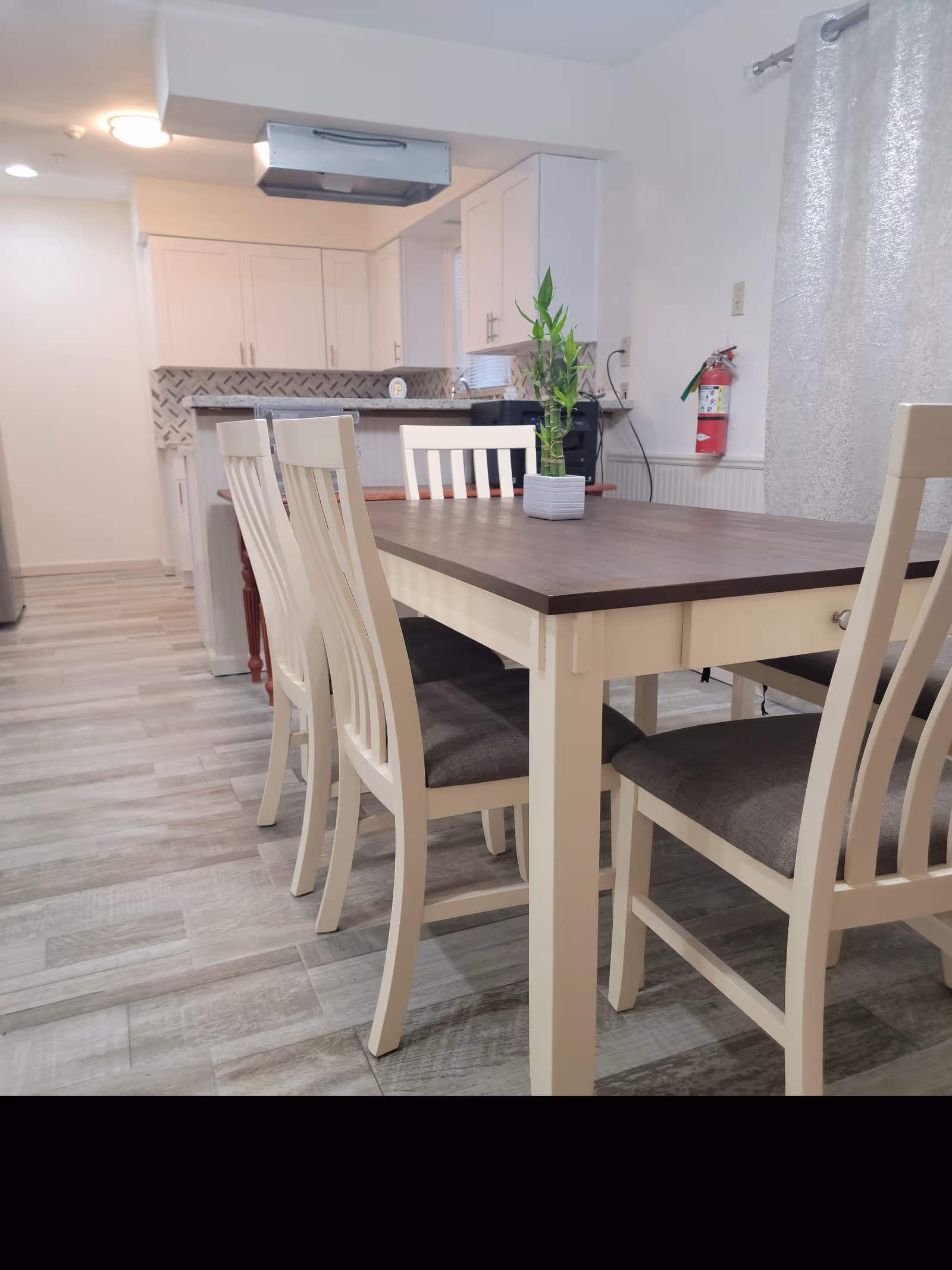 A modern dining area with a wooden table and six white chairs with dark cushions. A small potted plant is placed in the center of the table. In the background, there is a kitchen with white cabinets, a tiled backsplash, and a stainless steel range hood. The floor has light-colored tiles, and a fire extinguisher is mounted on the wall near a window with silver curtains.