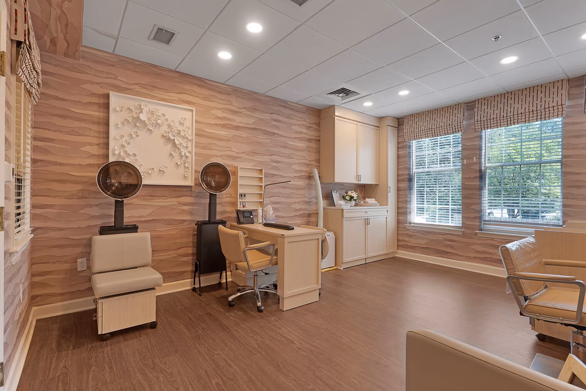 A well-lit salon room with two vintage hair dryers, a beige salon chair, a desk with a rolling chair, and cabinetry. The walls have a textured wave pattern wallpaper, and two large windows with blinds allow natural light to enter the room. The floor is wood, and the ceiling has recessed lighting.
