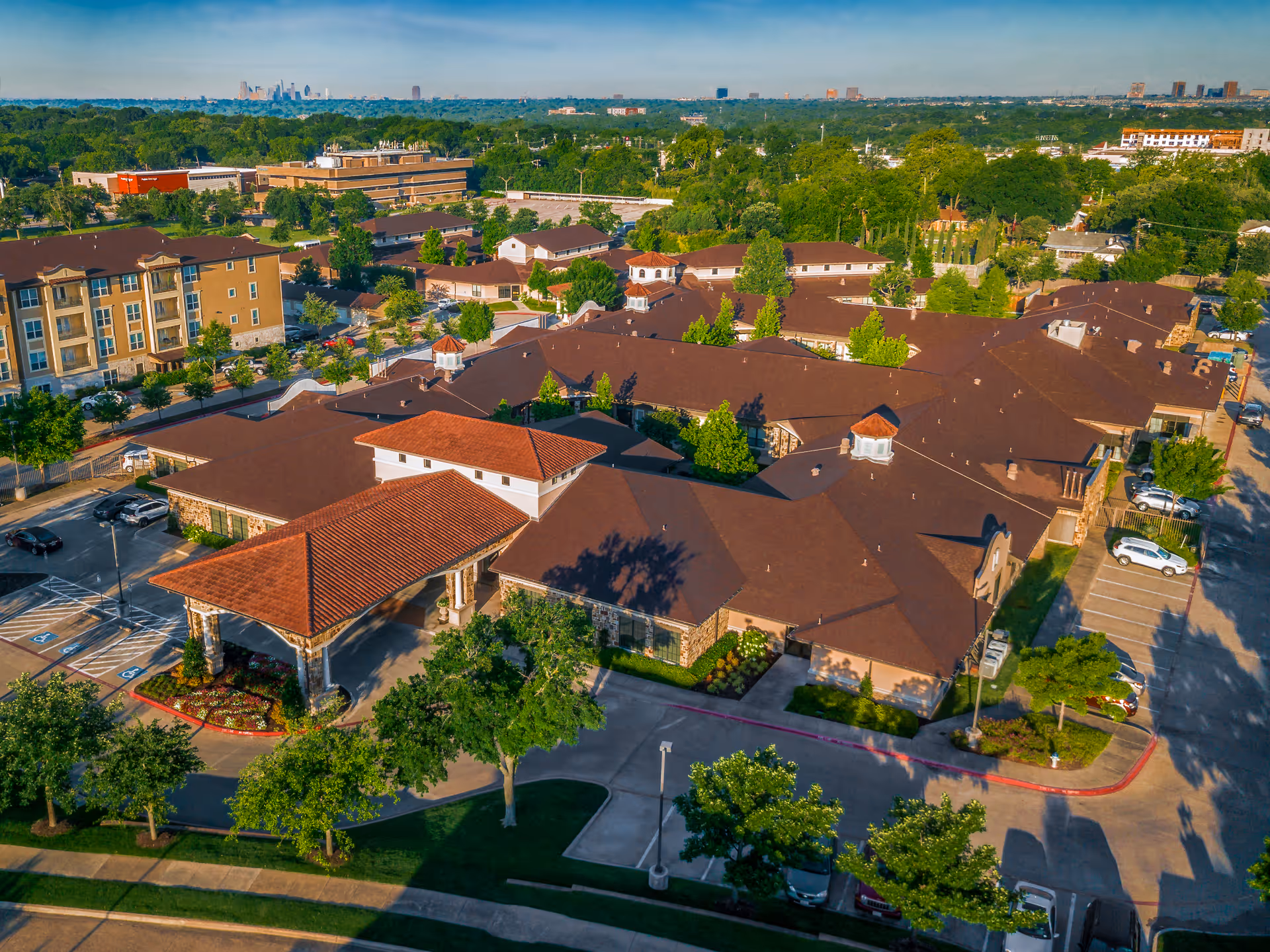 Aerial view of The Villages of Lake Highlands senior living facility showing multiple connected buildings with brown roofs, surrounded by trees, parking lots, and nearby residential and commercial buildings under a clear sky.