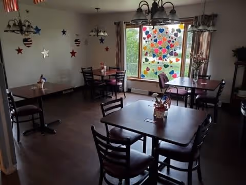 A dining room with several dark wooden tables and chairs arranged neatly. The room has a large window decorated with colorful heart-shaped cutouts. There are patriotic star decorations on the walls and small centerpieces on each table. The lighting fixtures hang from the ceiling, and curtains frame the window.