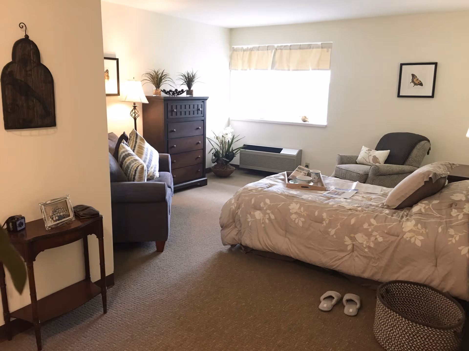 A cozy bedroom in a senior living facility featuring a bed with floral bedding, a cushioned armchair, a dark wooden dresser, a small sofa with striped pillows, a side table with a framed photo and a camera, and a window with a white valance letting in natural light. There are slippers on the carpeted floor and a woven basket near the bed.