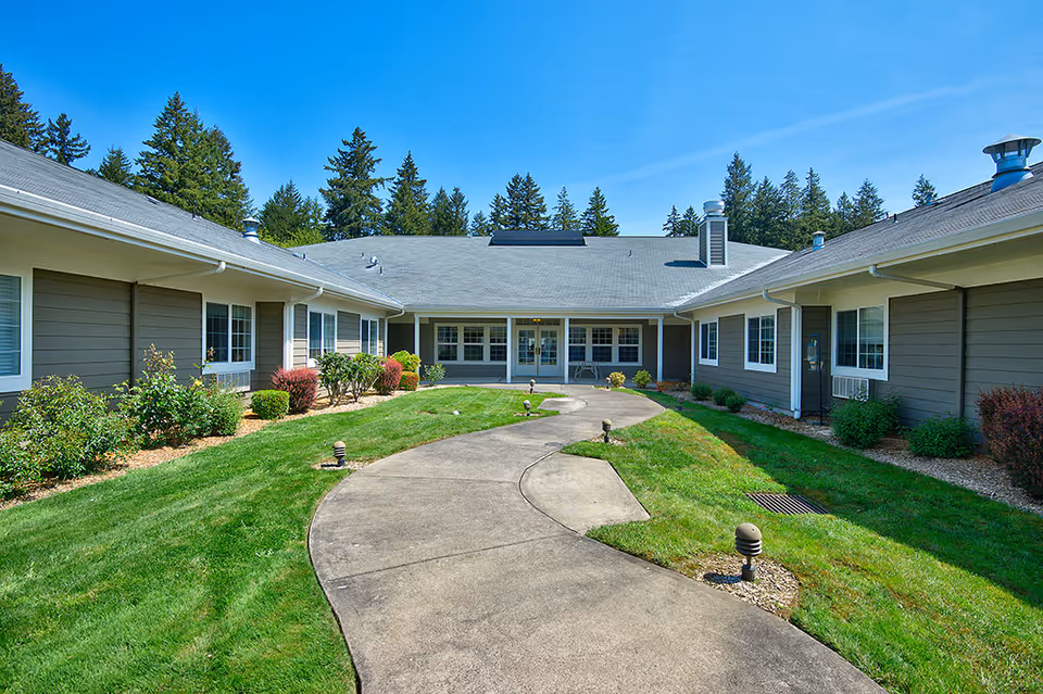 Curved concrete walkway leading through a landscaped courtyard to the entrance of a single-story retirement community building under a blue sky.