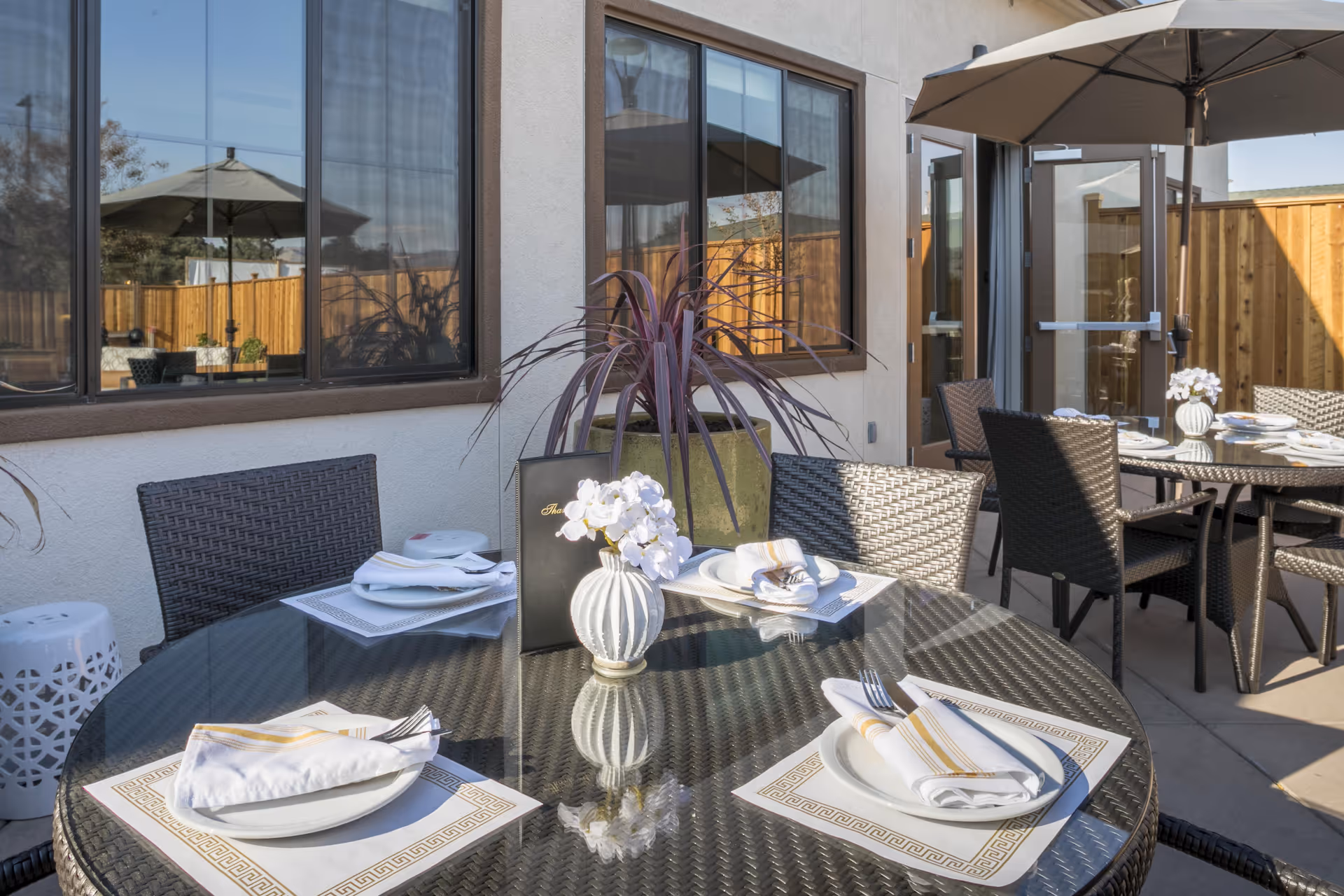 Outdoor patio area with round glass-top tables set for dining, each with white plates, folded napkins, and silverware. Wicker chairs surround the tables, and large umbrellas provide shade. A potted plant and a wooden fence are visible in the background.