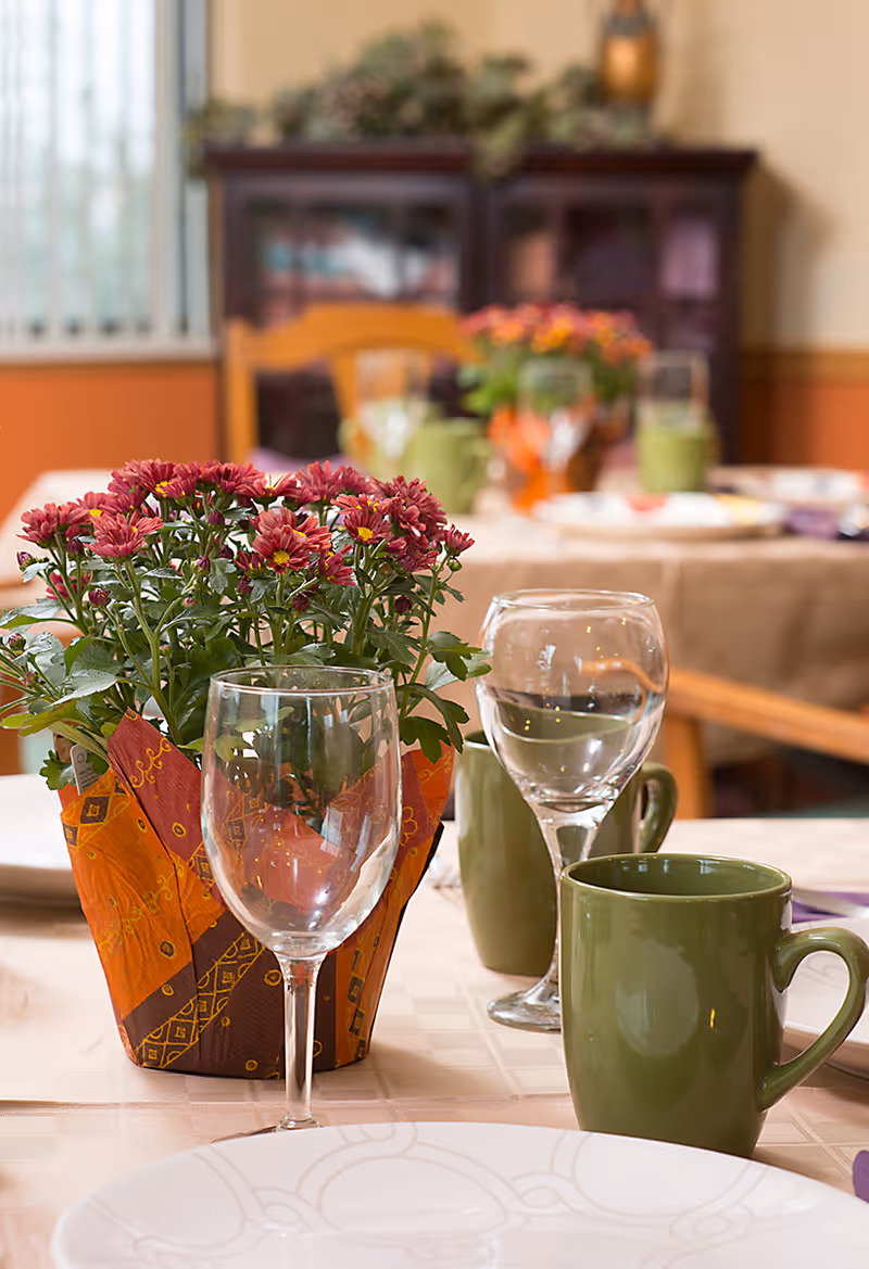 A dining table set with white plates, green mugs, and empty wine glasses. A small potted plant with red flowers wrapped in decorative orange and brown paper is placed in the center of the table. In the background, there are more tables similarly set and a wooden cabinet against a beige and orange wall.