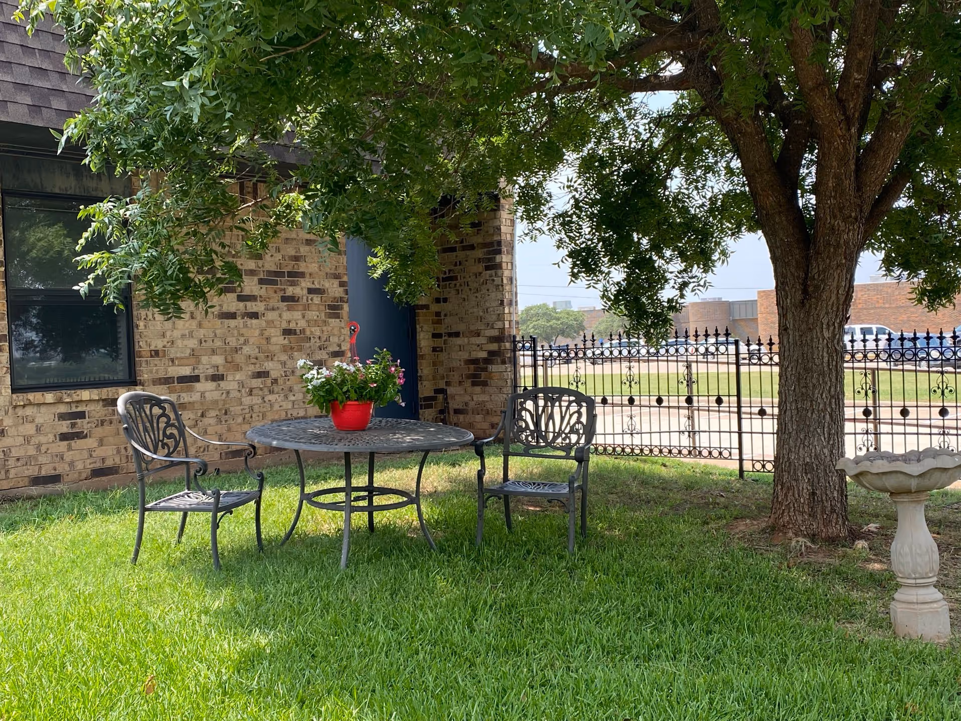 Shaded outdoor seating area with a metal table, two chairs and a potted plant under a tree beside a brick building and decorative fence.
