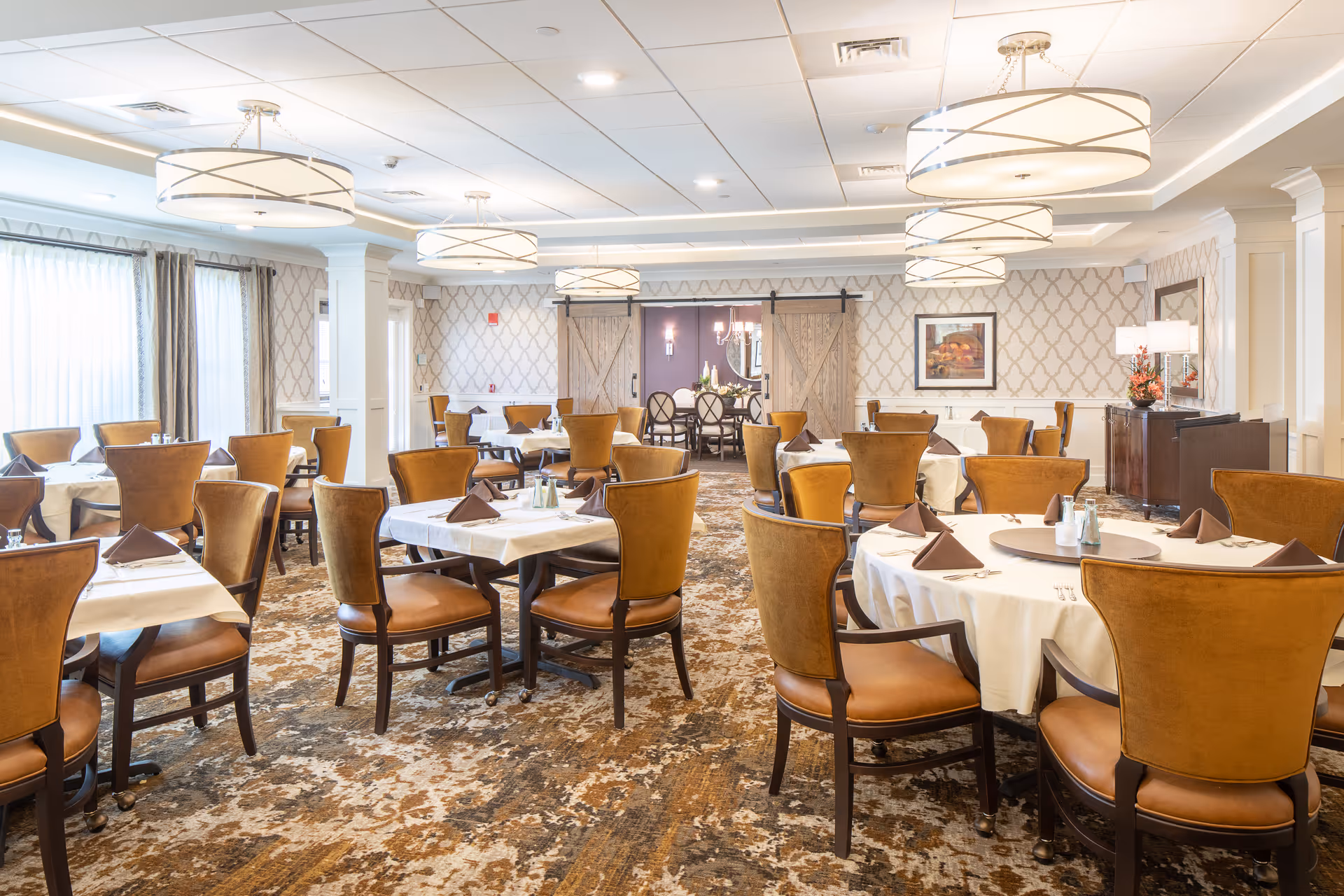 A spacious dining room with multiple round tables covered with white tablecloths and set with brown napkins, silverware, and condiments. The room features brown upholstered chairs, patterned carpet, large windows with curtains, and modern circular ceiling lights. In the background, there are sliding barn doors leading to another room with a chandelier and additional seating.