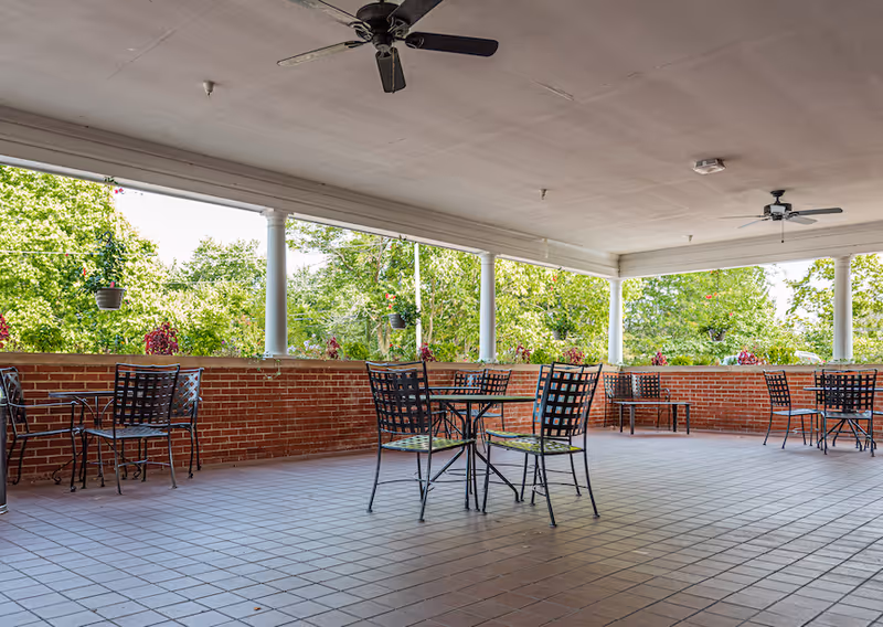 Covered outdoor patio with metal tables and chairs, brick half-walls, ceiling fans, and hanging plants overlooking trees.
