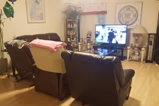 Cozy living room with recliner chairs and a loveseat arranged around a TV, with decorative shelves, wall art, and a floor lamp.