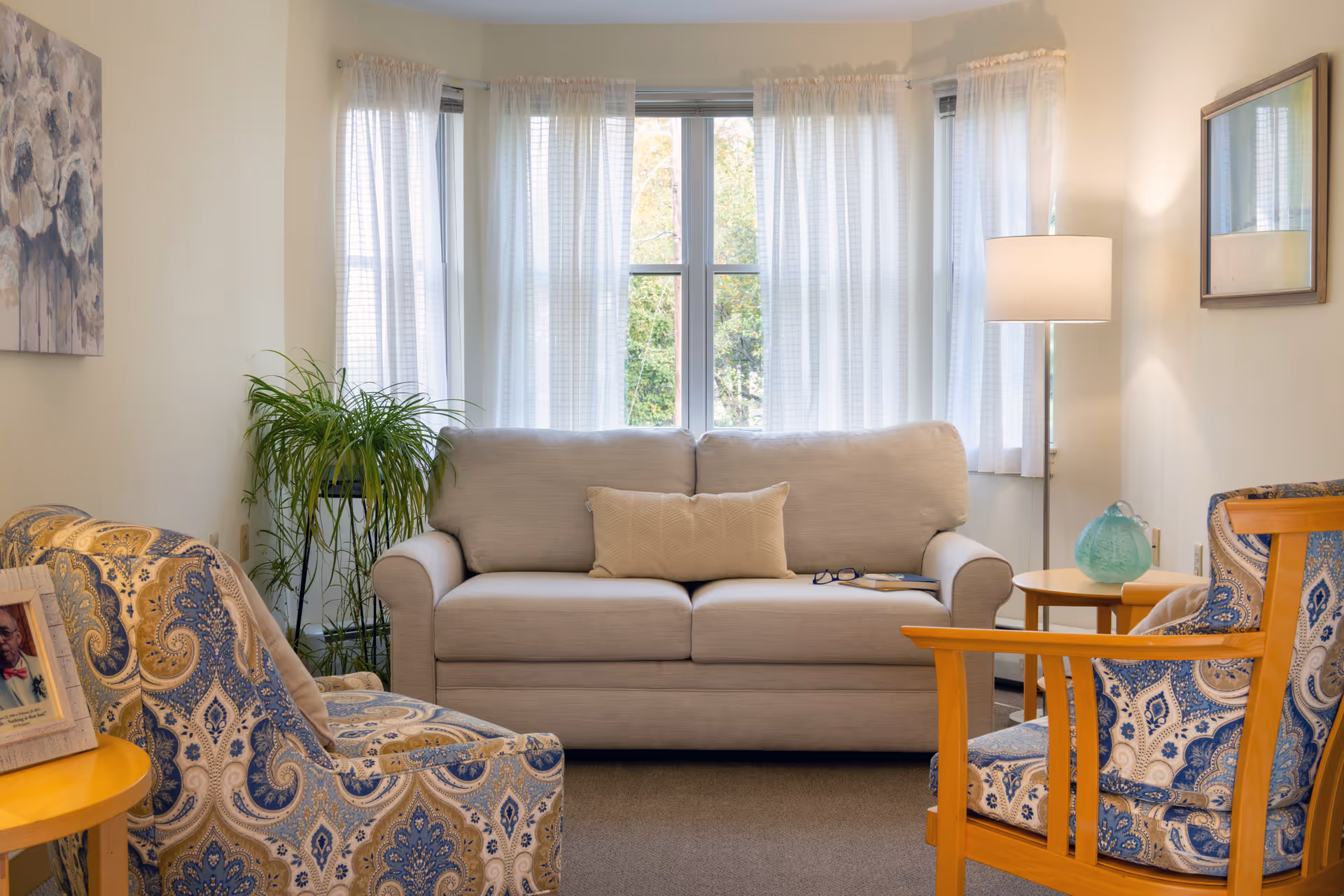 A cozy living room with a beige sofa centered in front of a window with sheer white curtains. Two patterned armchairs with blue and beige designs face the sofa. A small wooden side table with a green decorative glass object and a floor lamp with a white shade are on the right side. A potted plant is placed on a stand to the left of the sofa, and a framed photo and floral painting decorate the left wall.