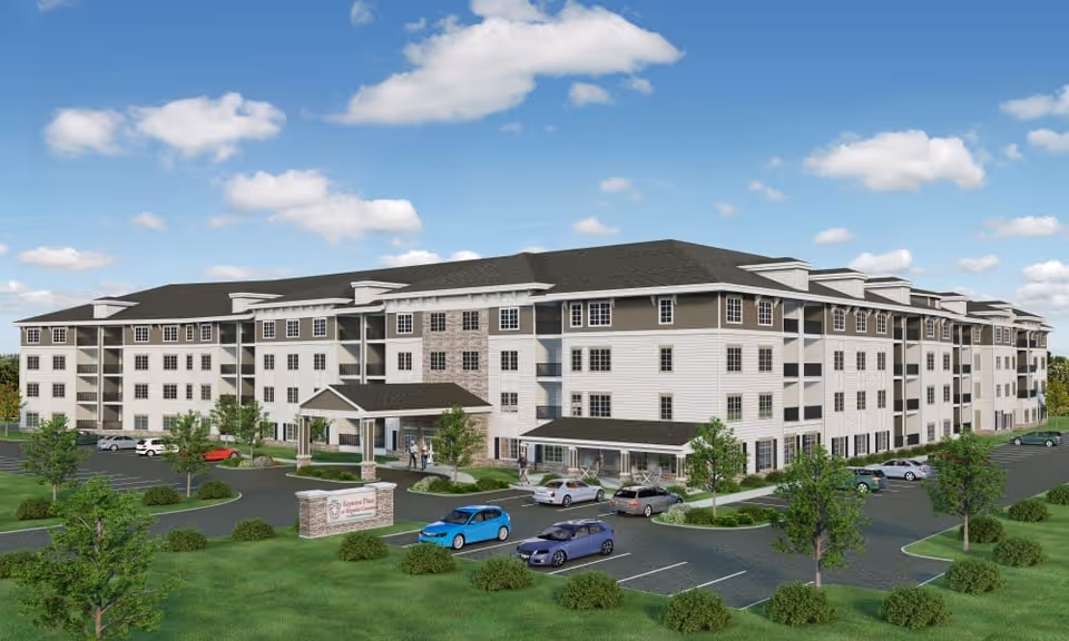 Exterior view of a large, multi-story senior living facility building with a covered entrance, multiple windows, and a parking lot with several cars. The building is surrounded by green lawns, trees, and bushes under a partly cloudy blue sky.