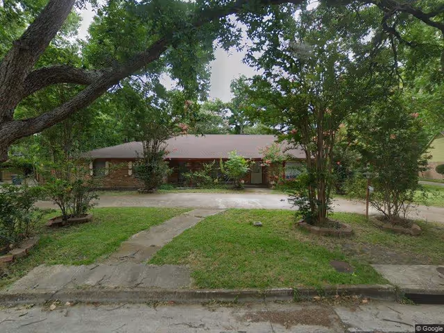 Single-story brick house with a brown roof surrounded by trees and greenery. There is a concrete driveway and a walkway leading to the front door. The yard has grass and several bushes and small trees.