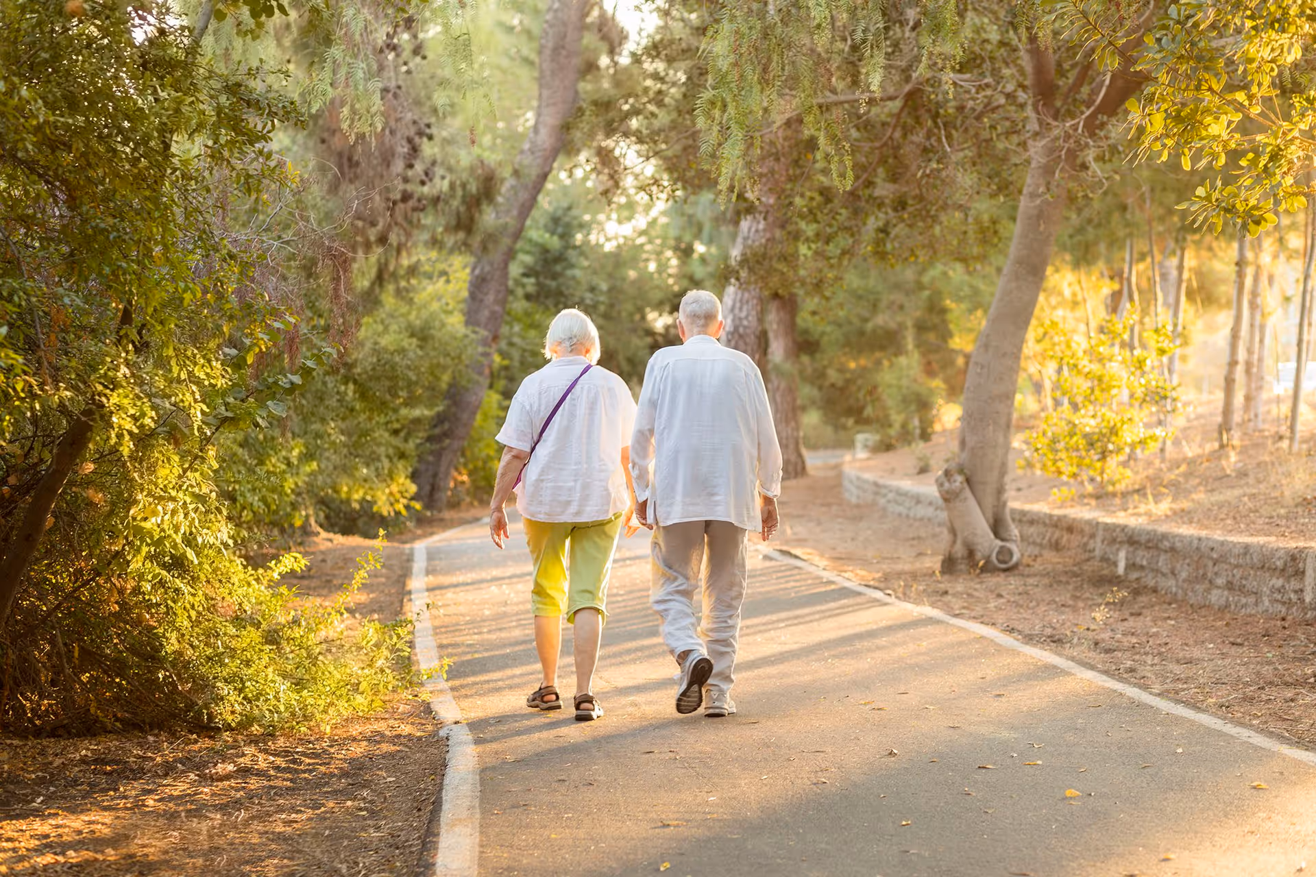 An elderly couple walking hand in hand down a paved path surrounded by trees and greenery in a park-like setting during golden hour.