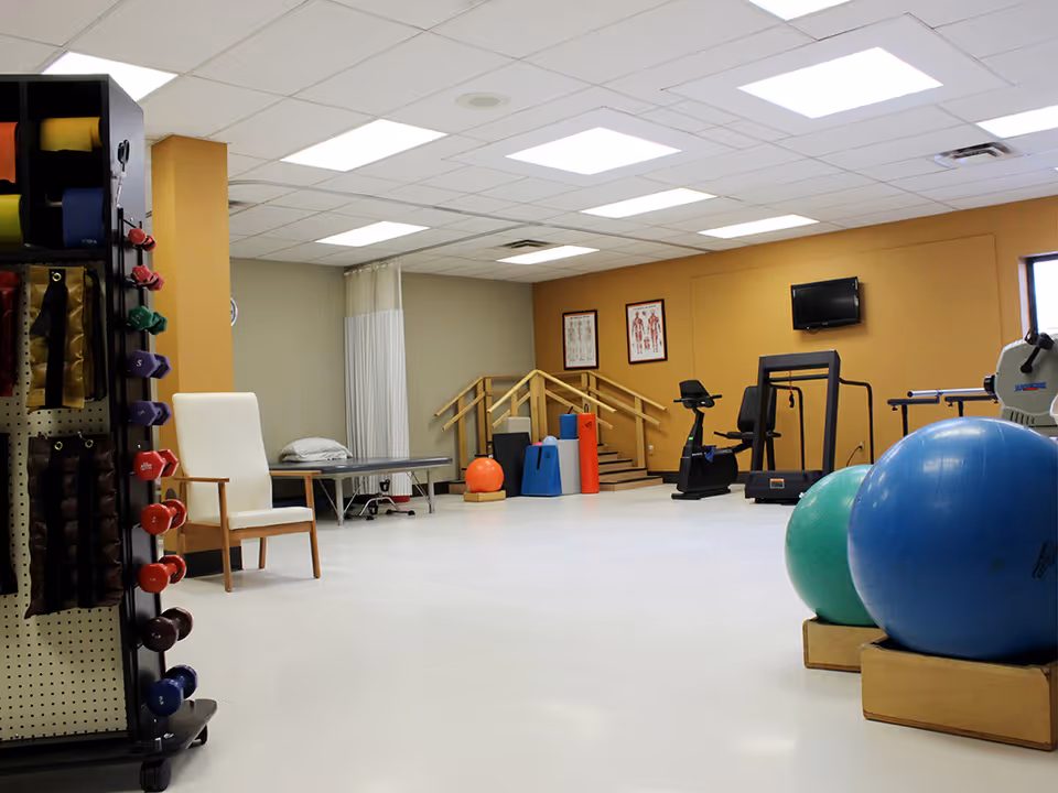 A spacious physical therapy or exercise room with various fitness equipment including exercise balls, stationary bikes, a treadmill, and weights. There is a white chair, a therapy table with a pillow, and anatomical charts on the wall. The room has a white ceiling with fluorescent lights and a beige and gray color scheme.