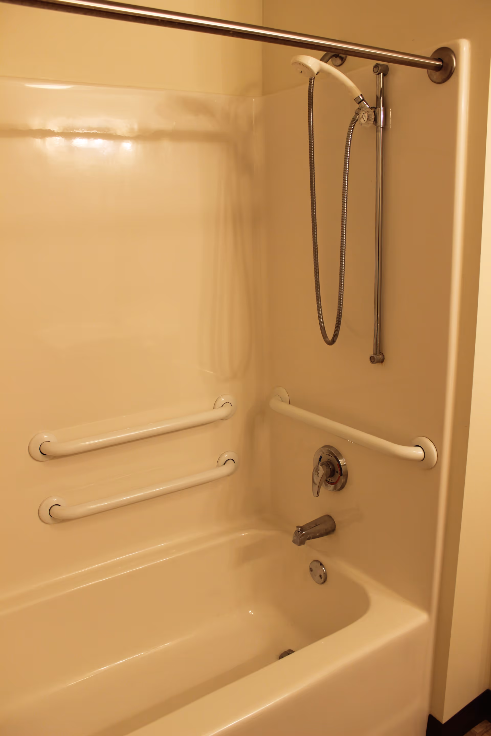 A beige bathtub and shower area with a handheld showerhead attached to a vertical sliding bar. The bathtub has three white grab bars installed on the walls for safety and accessibility.