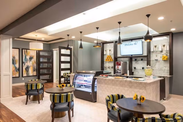A modern dining area in Heartis Mid Cities featuring round tables with striped cushioned chairs, a counter with a display case, beverage dispensers, and a wall-mounted screen displaying a welcome message. The room has pendant lighting, abstract artwork on the walls, and a warm, inviting atmosphere.