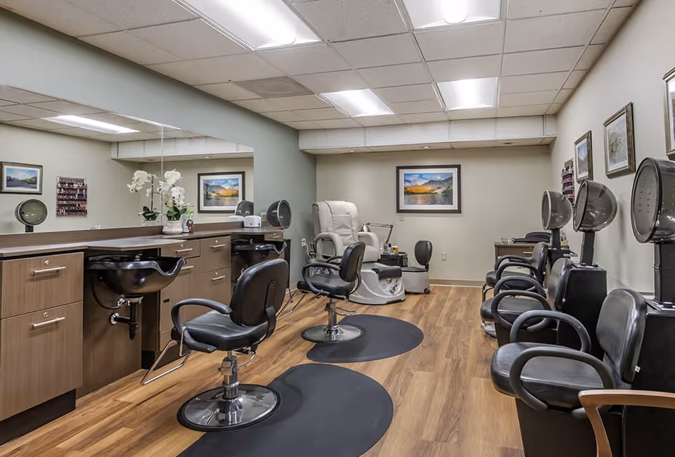 Interior view of a salon area in a senior living facility with multiple black salon chairs, hair drying stations, a pedicure chair, wooden flooring, and framed landscape pictures on the walls.