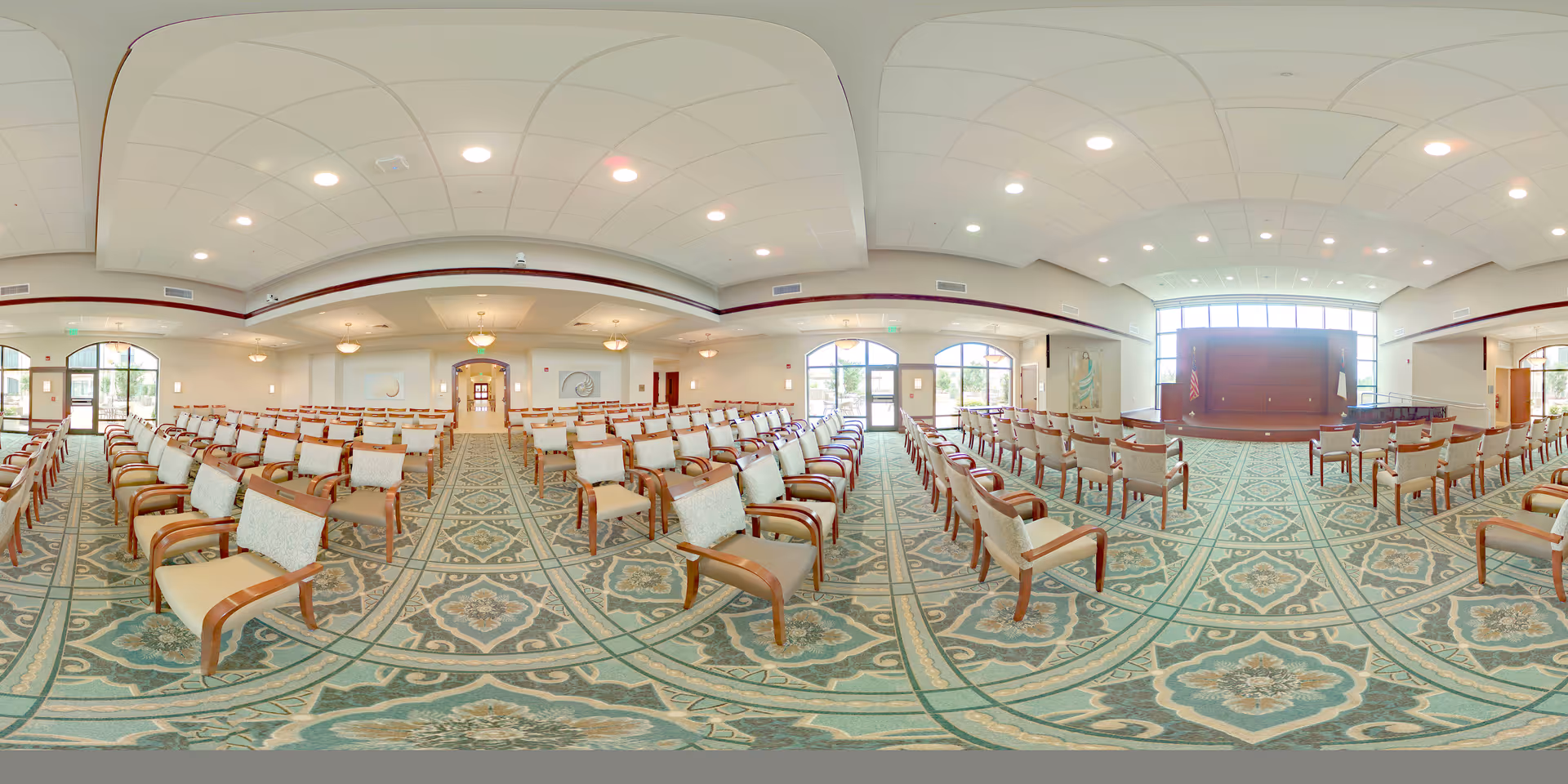 A large, well-lit meeting or event room with rows of beige cushioned chairs arranged facing a stage area with a podium. The room has large windows allowing natural light, patterned carpet flooring, and a high ceiling with recessed lighting. The walls are light-colored with some decorative elements and an American flag near the stage.