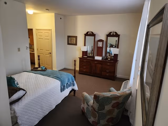 A bedroom in a senior living facility featuring a bed with a white quilt and teal throw, a patterned armchair, a wooden dresser with two mirrors, two table lamps, and a wall clock. There is a doorway leading to another room and a framed picture on the wall.