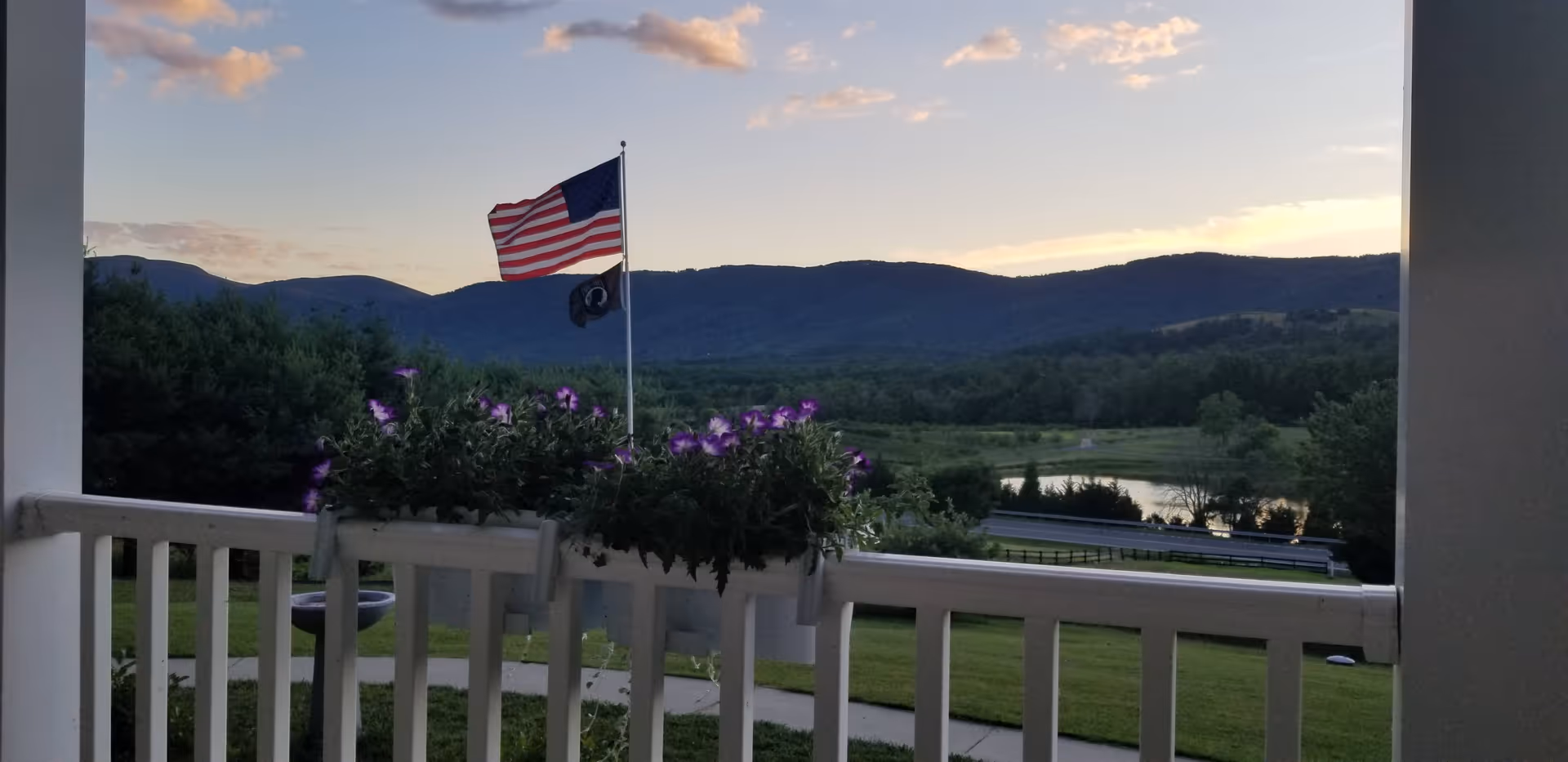 Balcony view with purple flowers, railing, flag, and distant mountains.