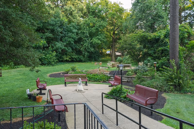 Outdoor garden courtyard with several red benches arranged around a small concrete patio and statue, surrounded by grass and trees.