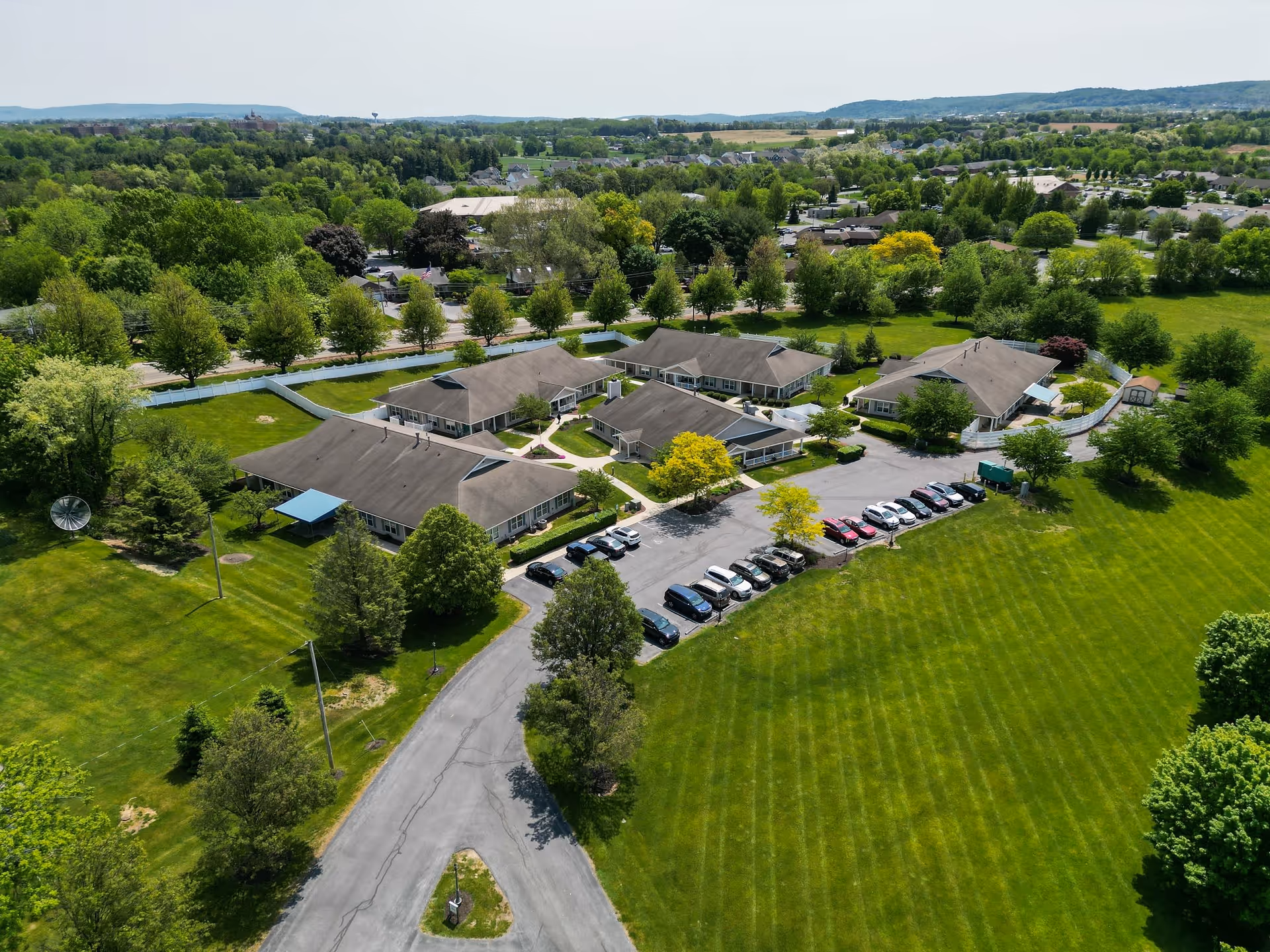 Aerial view of Linden Village A ProMedica Senior Living Community showing multiple single-story buildings surrounded by green lawns, trees, and a parking area with several cars. The facility is set in a suburban area with more greenery and houses in the background under a clear sky.