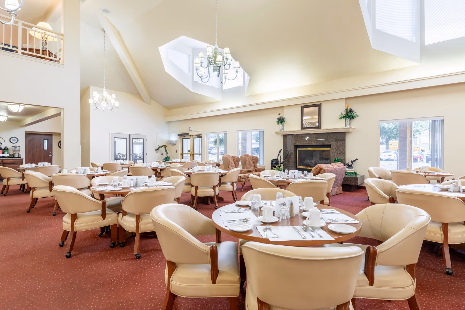 Spacious senior living dining room with round tables set for meals, beige chairs, chandeliers and a fireplace.