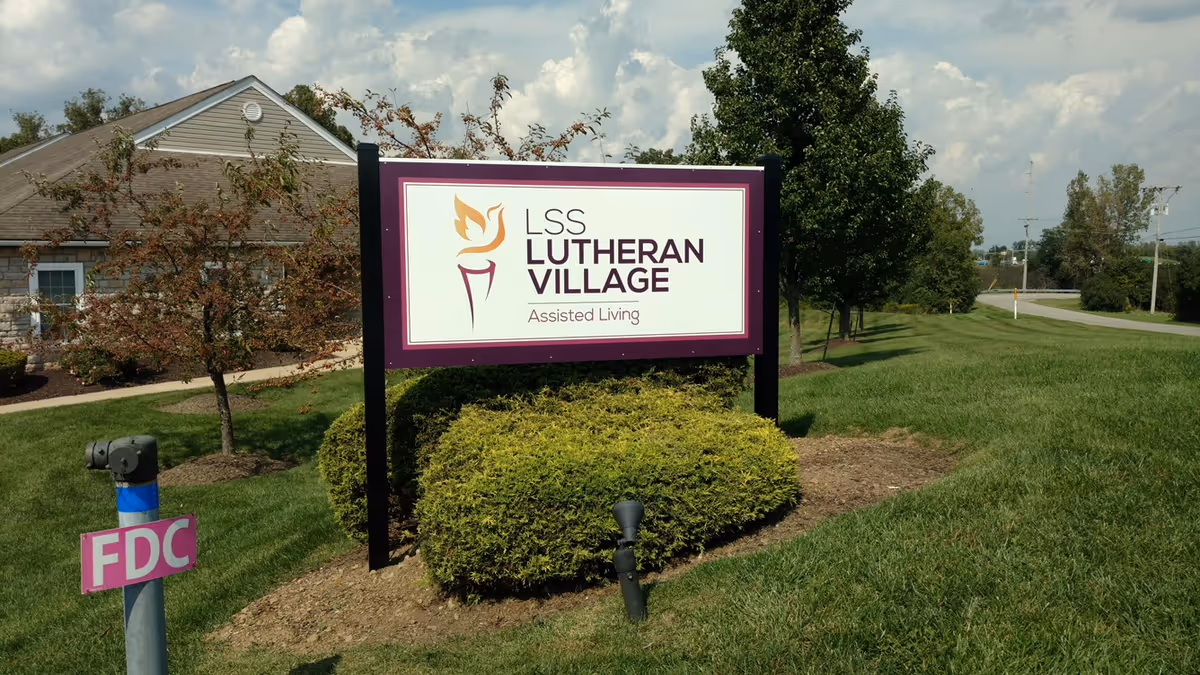 Outdoor view of a sign for LSS Lutheran Village Assisted Living, positioned on a grassy area with bushes and trees nearby. A building with a stone facade and a shingled roof is visible in the background under a partly cloudy sky.