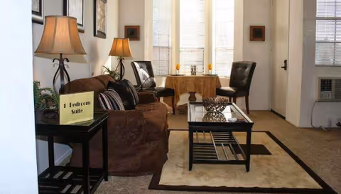 A cozy living room with a brown sofa, two black chairs, a glass coffee table on a beige and brown area rug, two table lamps on side tables, and a small table with a yellow sign that reads '1 Bedroom Suite'. The room has large windows with blinds and light-colored walls.